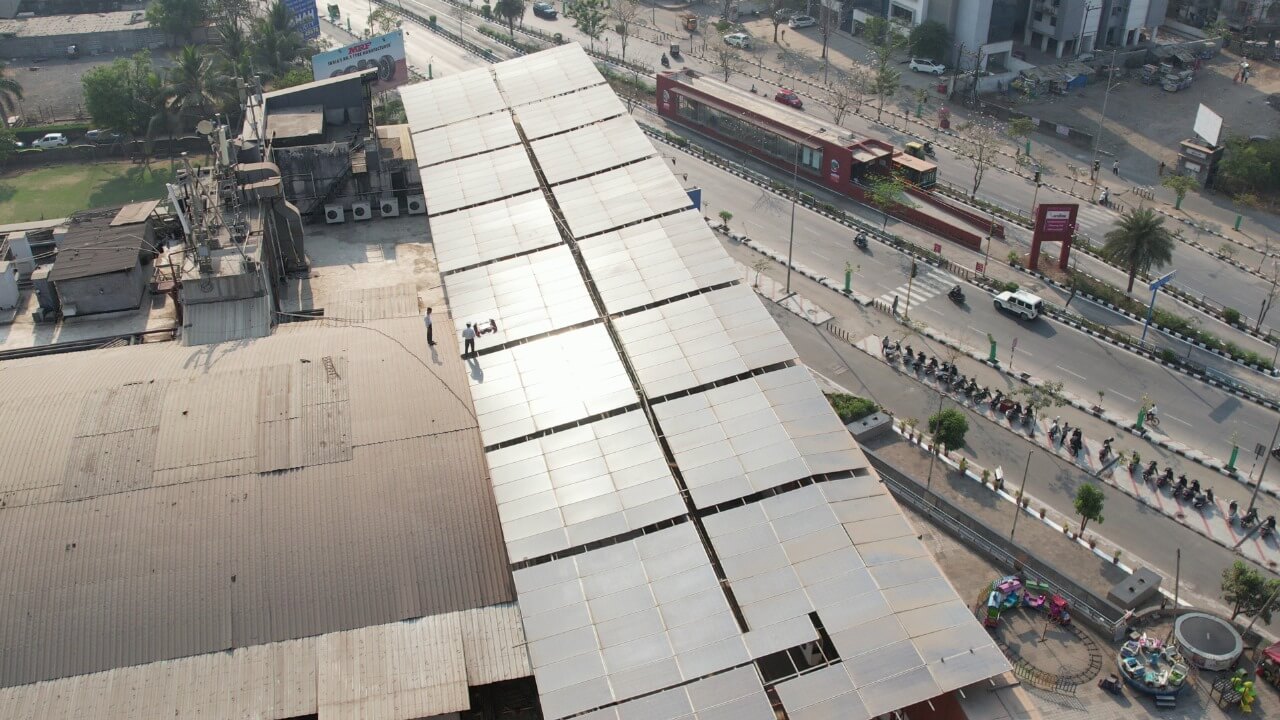 Two workers inspecting a long array of PV panels on a Rahul Raj commercial mall roof above a busy street